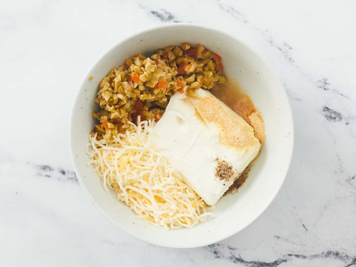 A small white bowl filled with unmixed ingredients of a cream cheese spread, including a block of cream cheese, shredded cheddar cheese, chopped pimento-stuffed olives and olive brine on a white marble background.