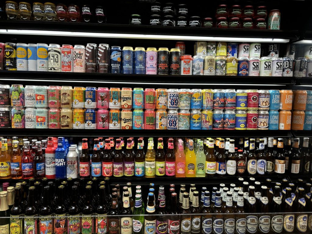 A well-stocked refrigerator display of various beers and alcoholic beverages in cans and bottles. The shelves
